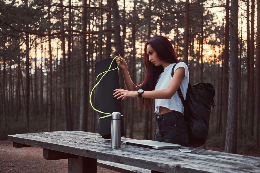 Une jeune femme ajuste un tapis de sol à côté d'une gourde filtrante nomade en inox posée sur une table en bois dans une forêt de pins.