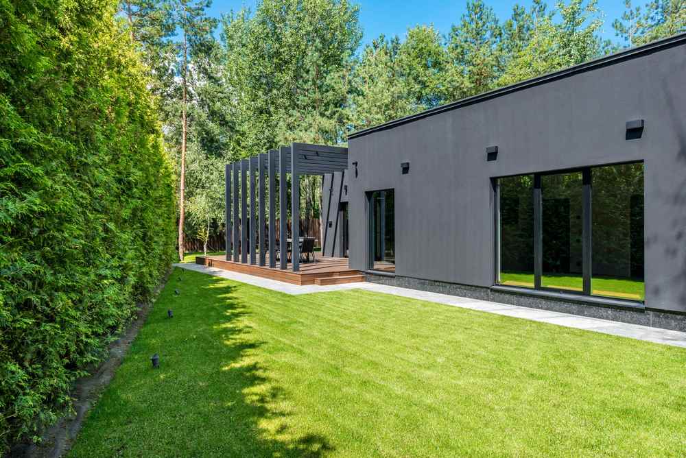 Maison gris avec grandes fenêtres, terrasse en bois, pelouse bien entretenue, arrière-jardin arboré